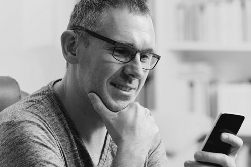 A middle aged man wearing glasses is holding a smart phone, looking at it and smiling slightly.